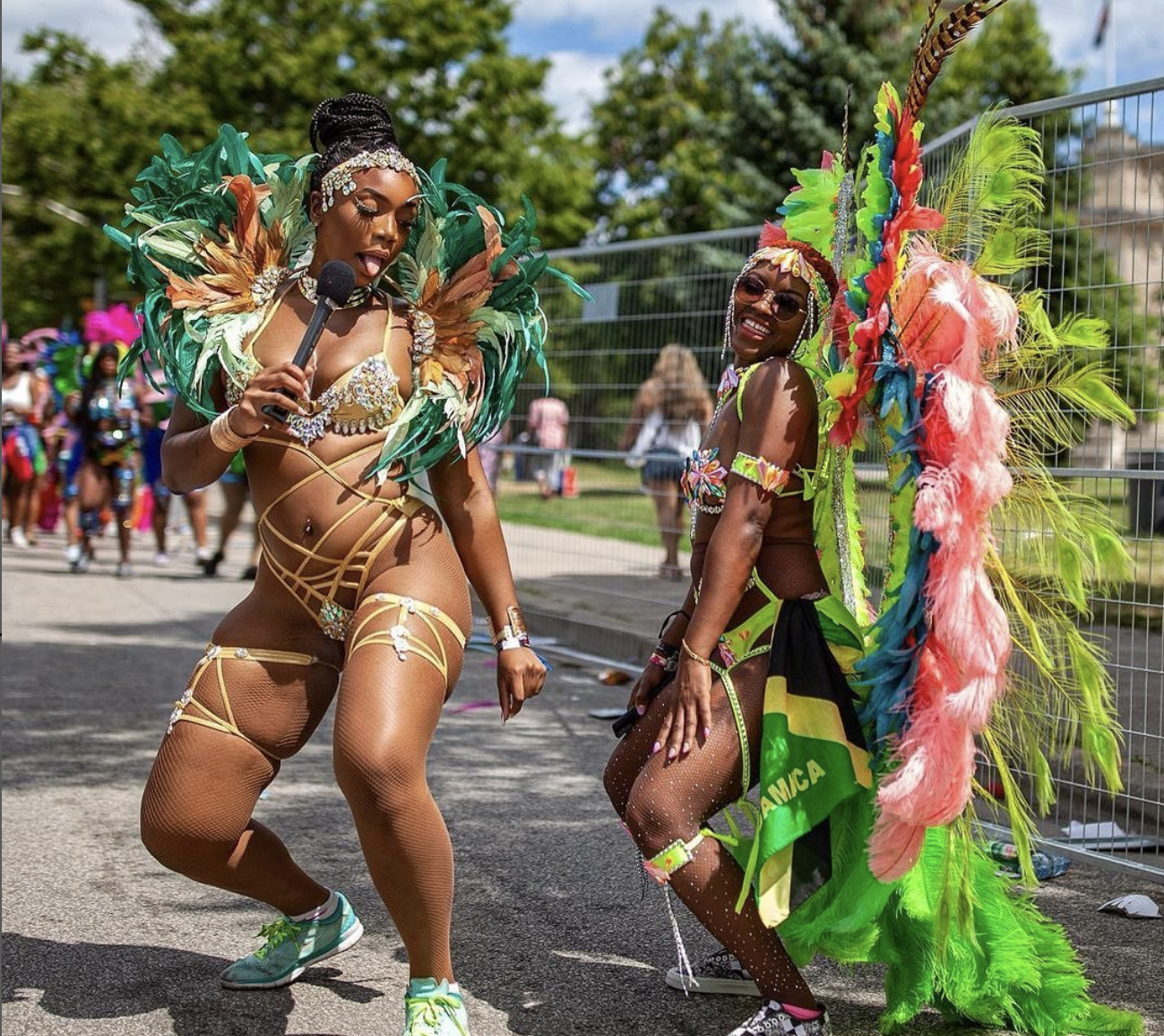 Two Sunlime Mas masqueraders at the 2022 Toronto Caribbean Carnival. One masquerader is dawning a green feathered costume while interviewing a second female masquerader dawning a large yellow feathered frontline costume.